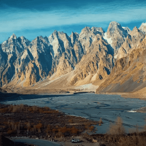 Wide-angle landscape view of the jagged rock spires of Passu Cones (Tupopdan Peaks) under a bright blue sky with light clouds, overlooking the dry river bed and Karakoram Highway curve in Gojal, Upper Hunza Pakistan.
