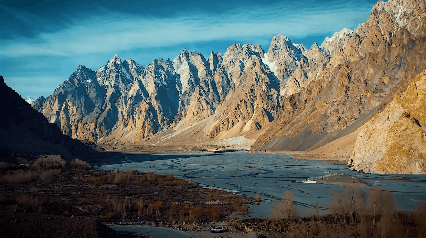 Wide-angle landscape view of the jagged rock spires of Passu Cones (Tupopdan Peaks) under a bright blue sky with light clouds, overlooking the dry river bed and Karakoram Highway curve in Gojal, Upper Hunza Pakistan.