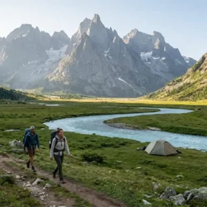 Two backpackers walking along a valley stream with a campsite tent and sharp granite mountain peaks in the background. Things to Do on a Mountain