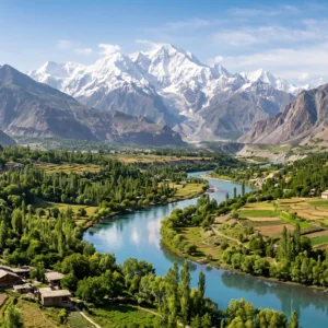 Aerial view of Ghizer Valley in northern Pakistan showing turquoise river winding through green fields and poplar trees with snow-capped Hindu Kush peaks in the background