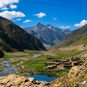Panoramic view of Batakundi valley in Kaghan showing a winding road, traditional stone houses, lush green terrace fields, and a rushing river stream with snow-capped mountains under a blue sky.