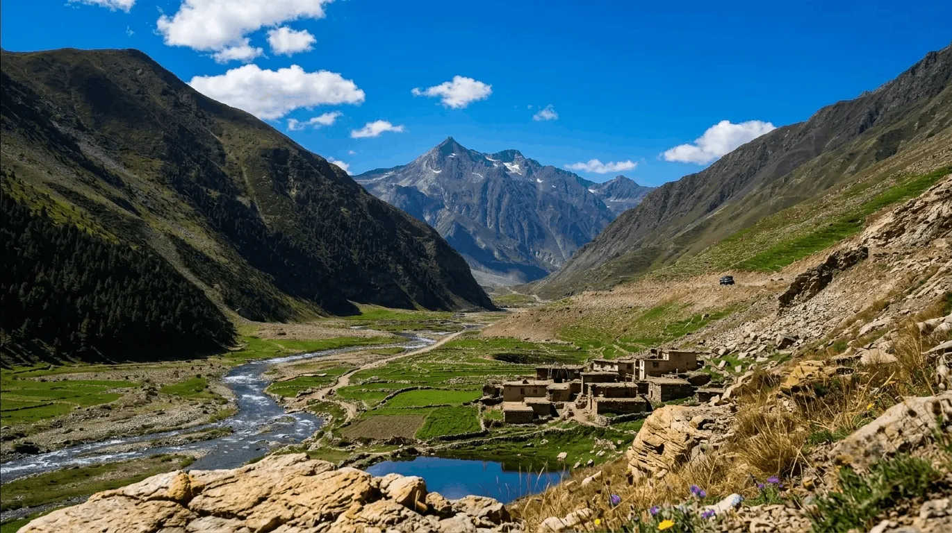 Panoramic view of Batakundi valley in Kaghan showing a winding road, traditional stone houses, lush green terrace fields, and a rushing river stream with snow-capped mountains under a blue sky.